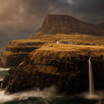 A dramatic landscape view of Mullafossur waterfall cascading into the ocean with Gasadalur village perched on the cliffs, under a cloudy sky in the Faroe Islands