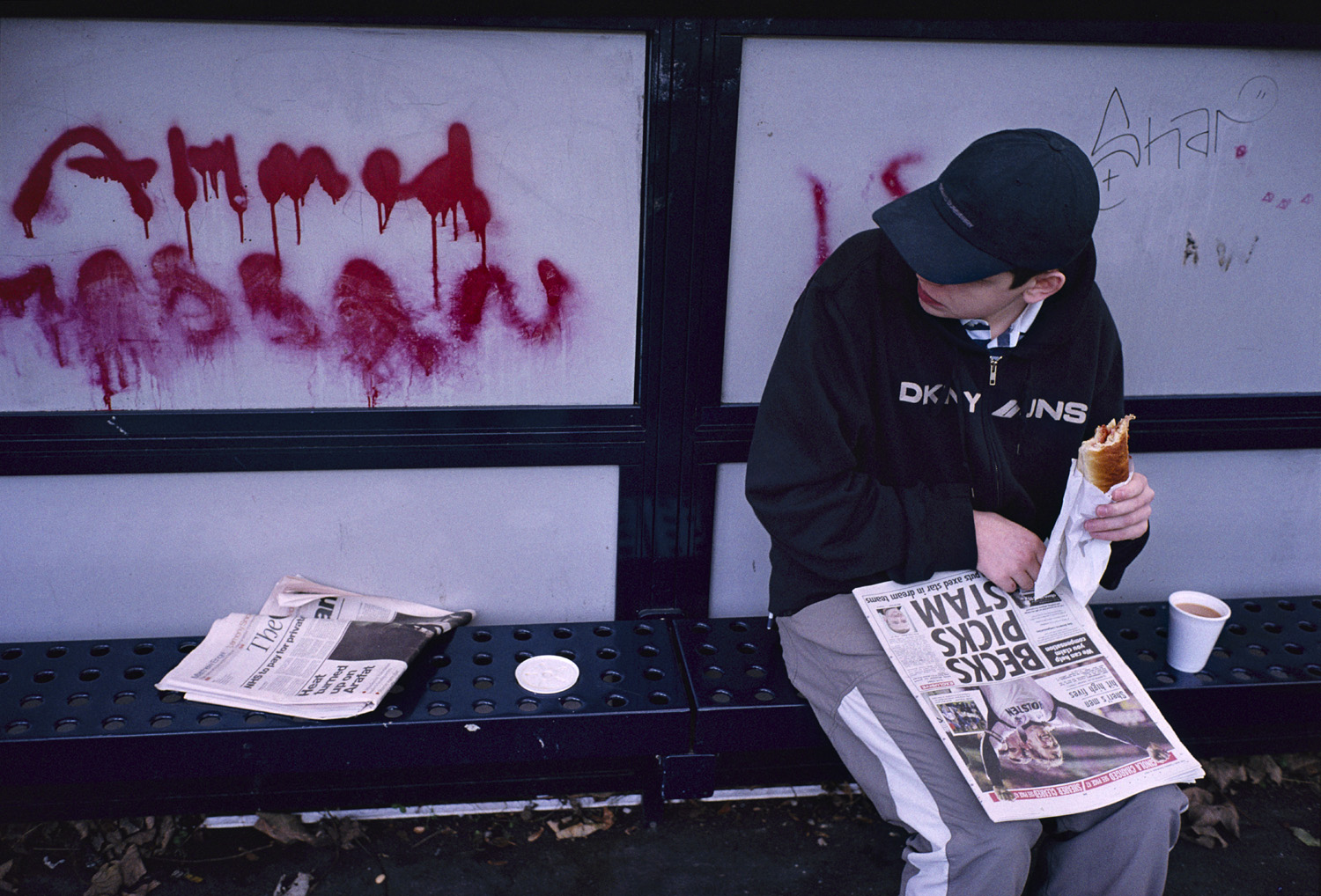 A young man sits on a bench at a train station, holding a bacon sandwich and reading a newspaper with graffiti on the wall behind him.