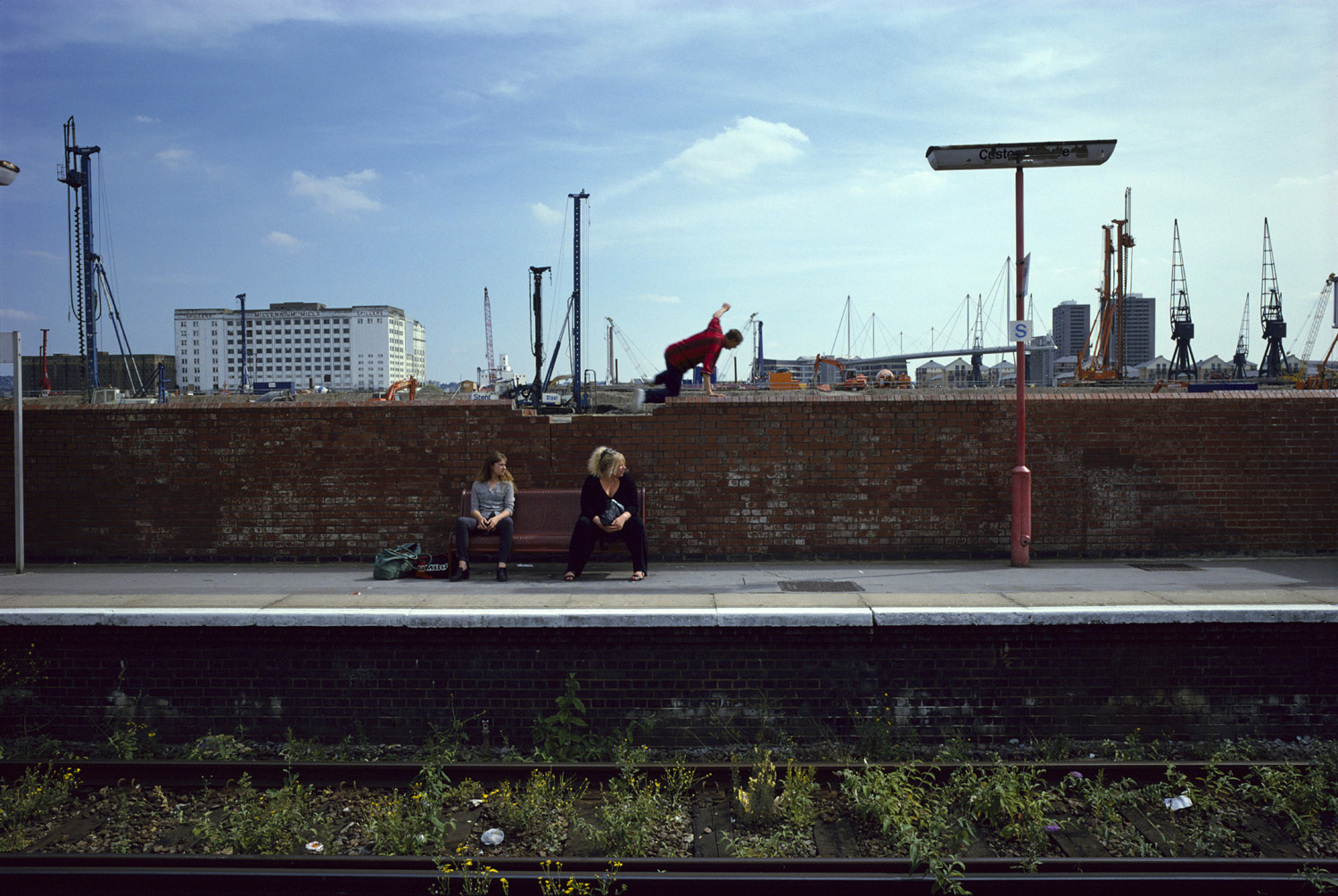 Two women sit on a bench at a train station platform with industrial cranes and buildings in the background, while a man in mid-air vaults over a brick wall.
