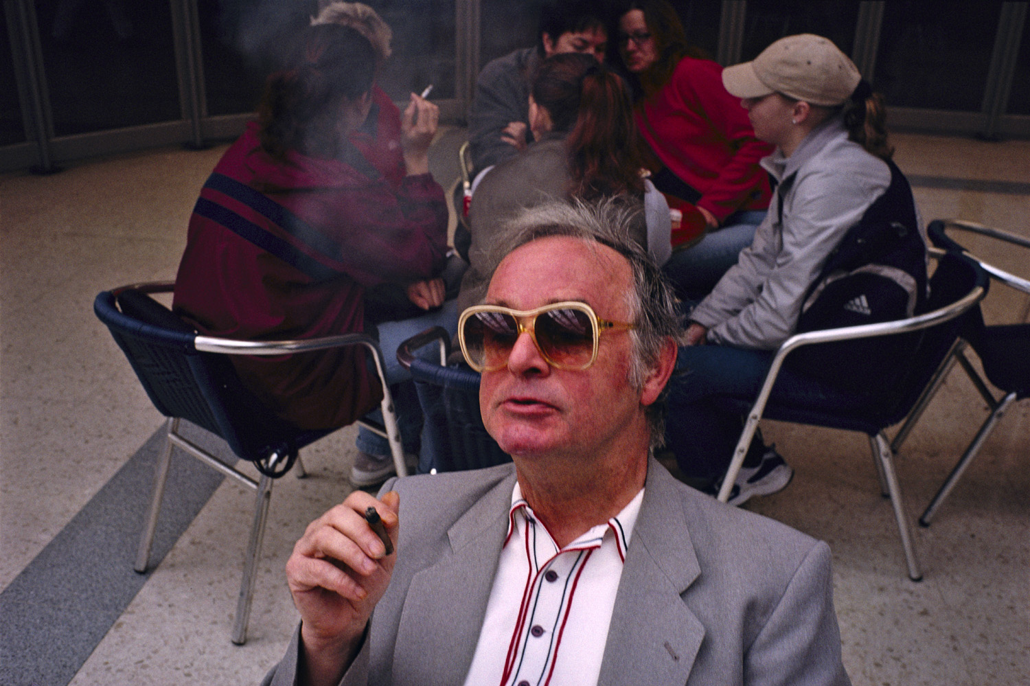 A grey-haired man in sunglasses and a suit smokes a cigarette, with a group of young people smoking and chatting in the background.