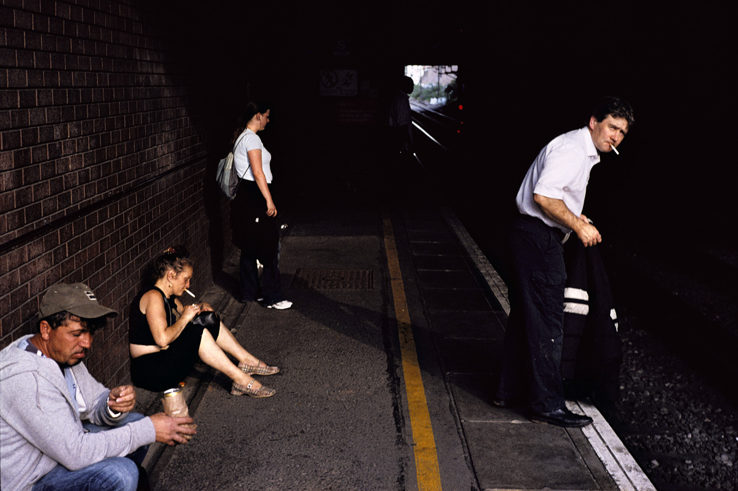 A group of people wait on a station platform in golden hour light, each absorbed in their own world, highlighting a moment of urban solitude.