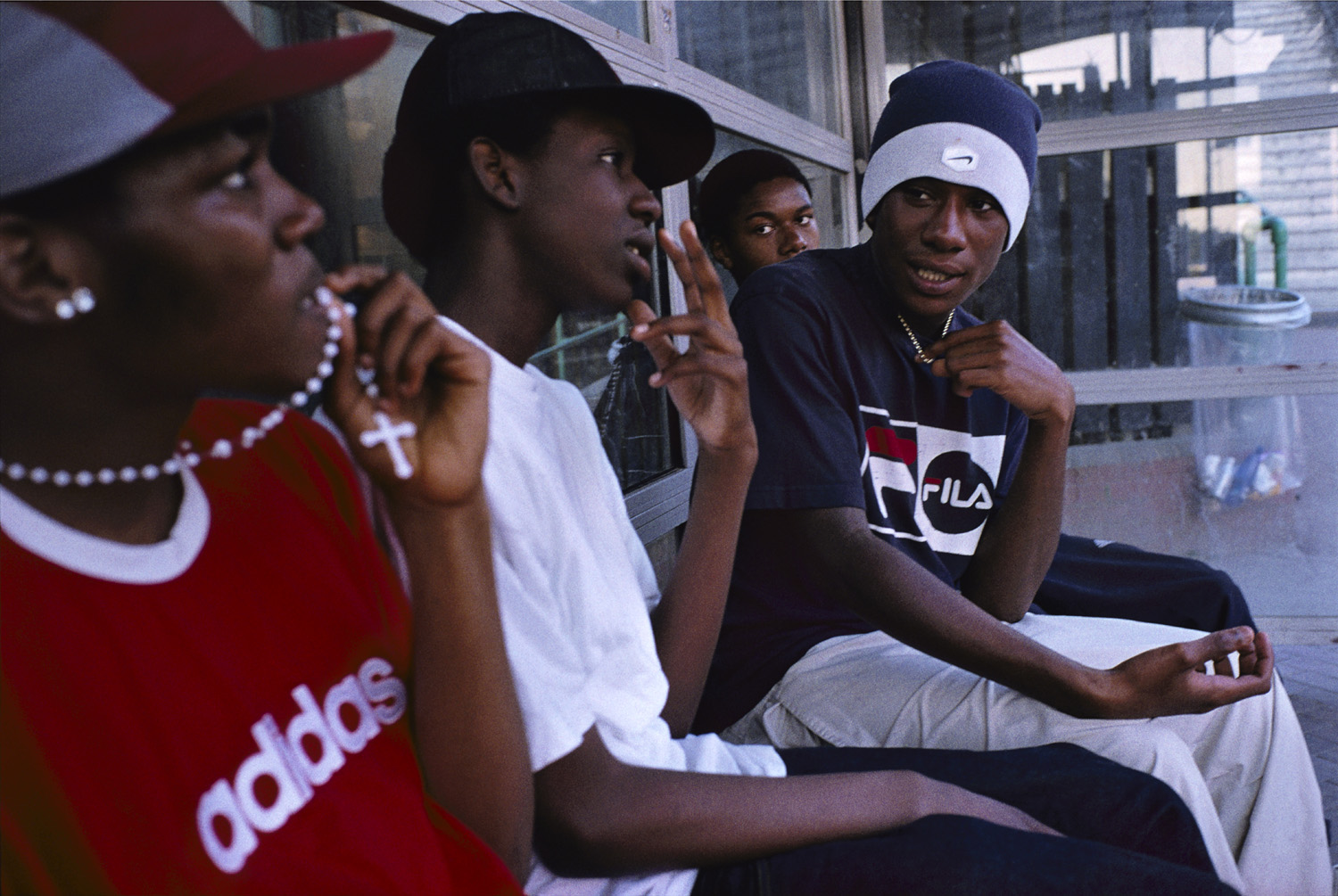 A group of young men in casual attire, including baseball caps and sportswear, sit and chat on seats at a train station, embodying youth culture.