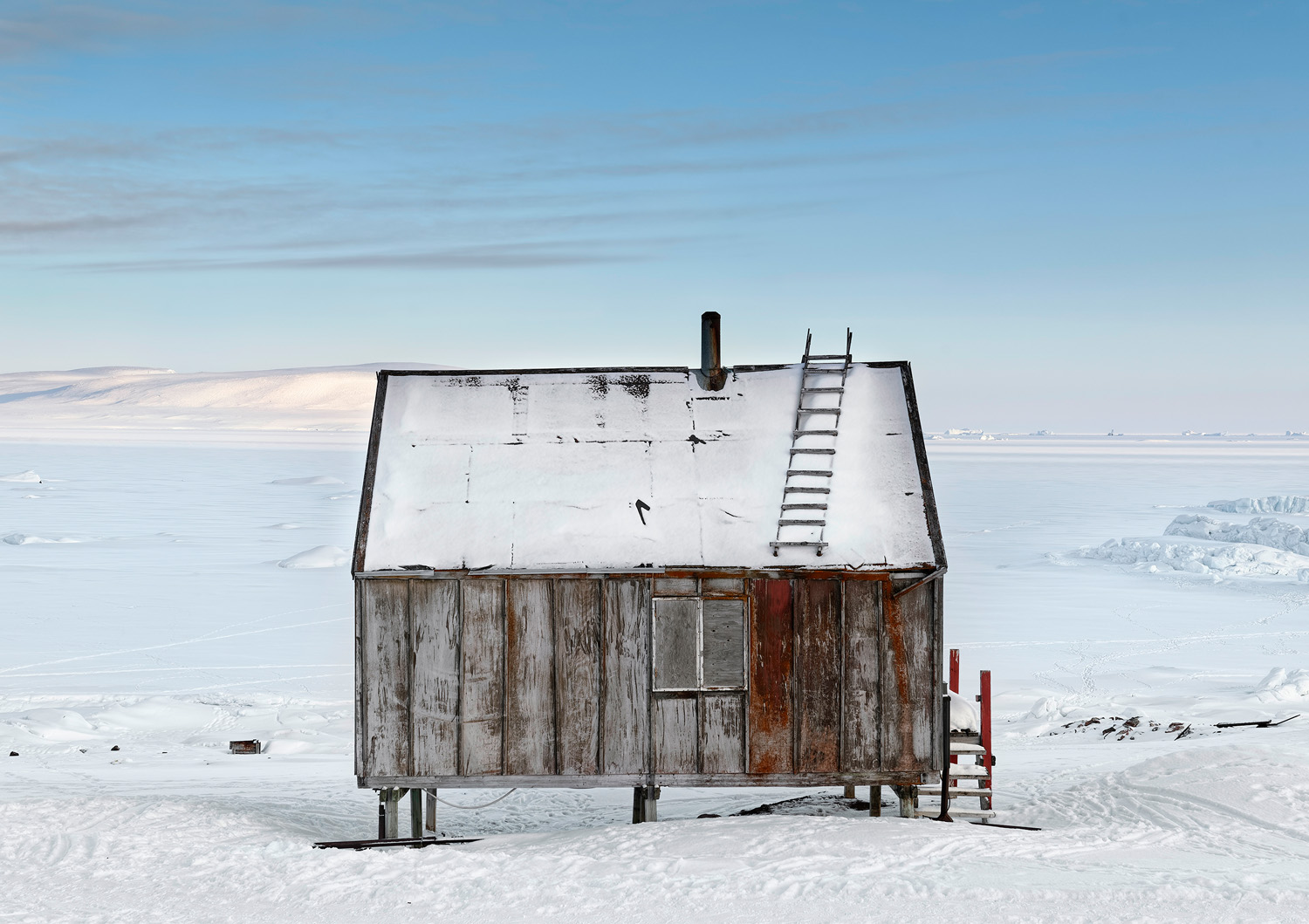 A solitary wooden cabin with a sloped roof covered in snow, set against a vast, snowy landscape under a clear blue sky.