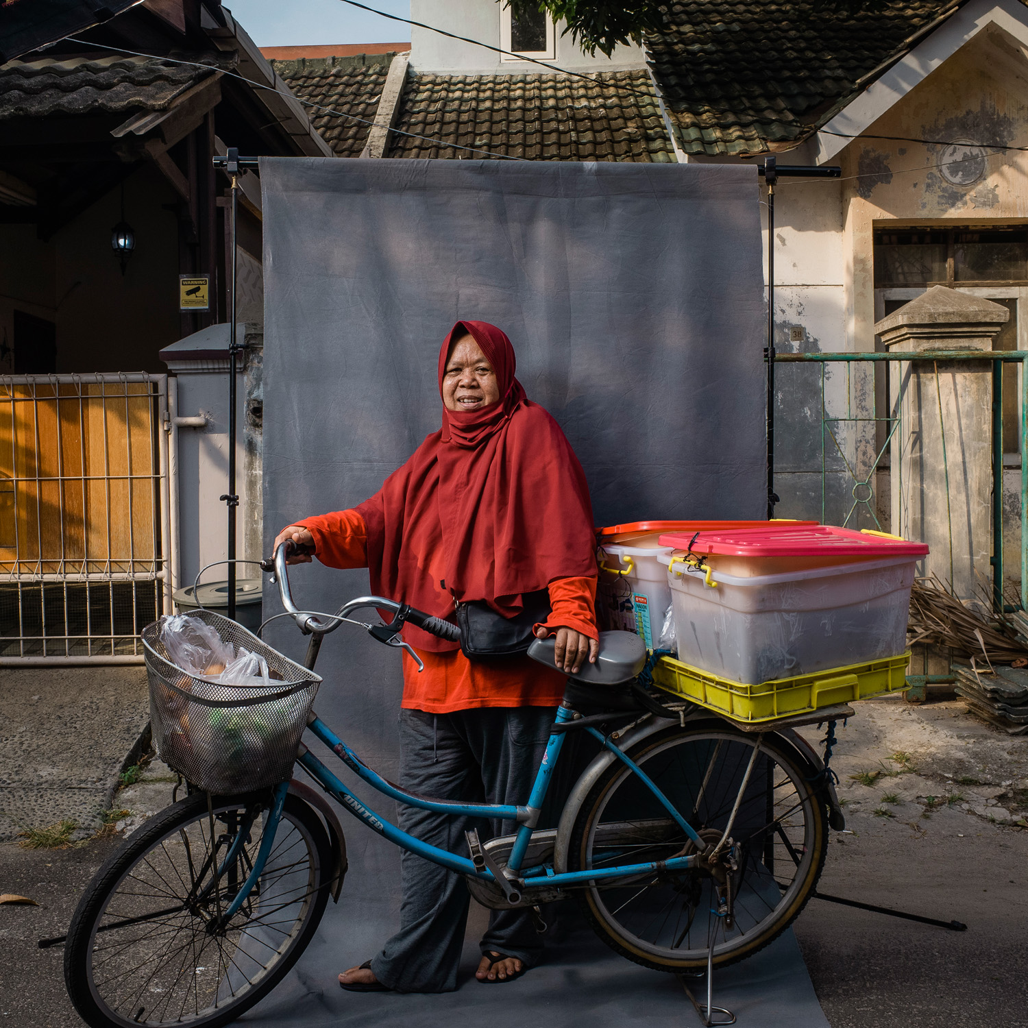 A woman wearing a red headscarf and an orange shirt stands beside her bicycle loaded with goods. She is in front of a grey backdrop set up on a street with houses in the background.