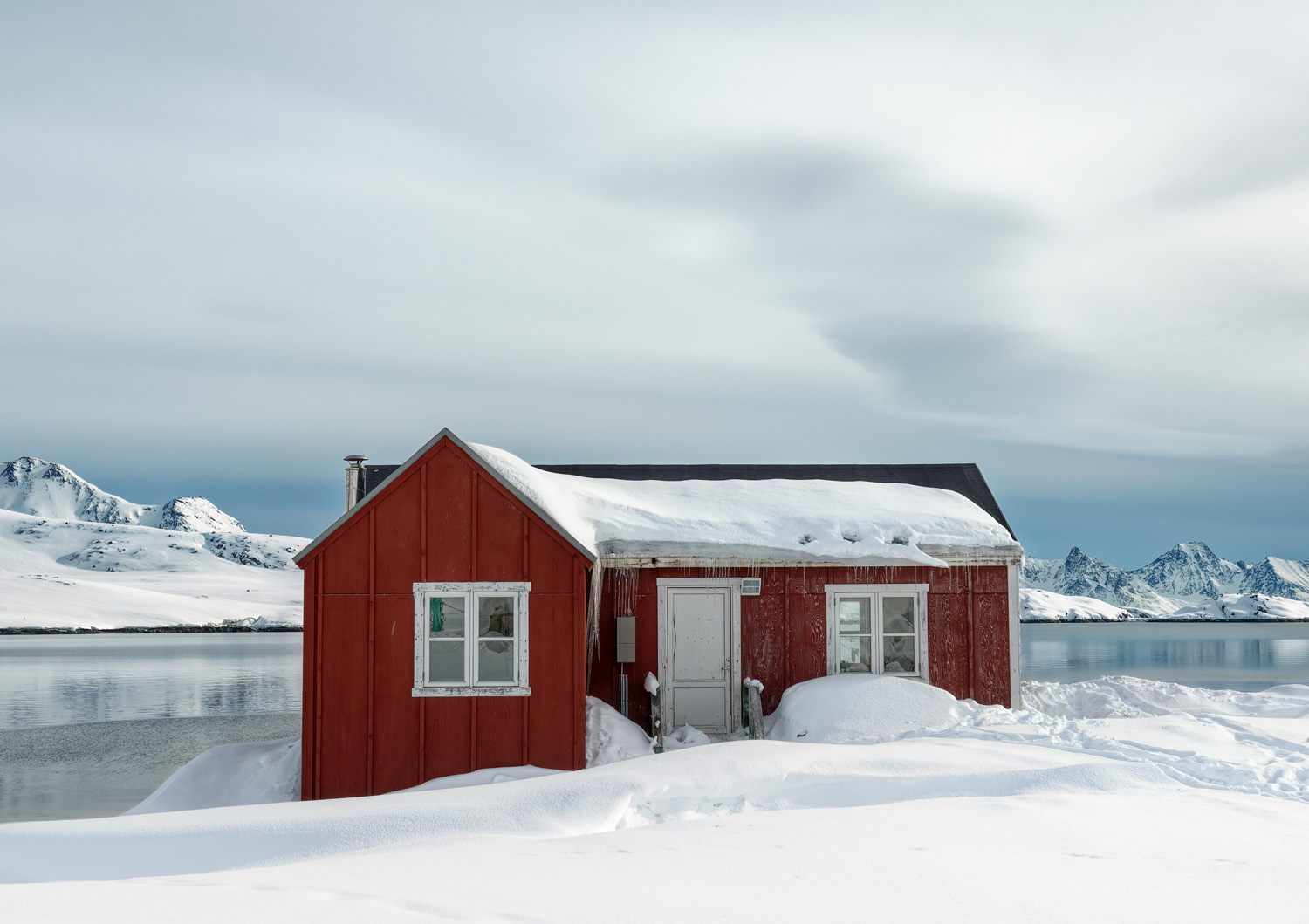 A red wooden house with a snow-covered roof situated near a calm lake with snow-capped mountains in the background under an overcast sky.