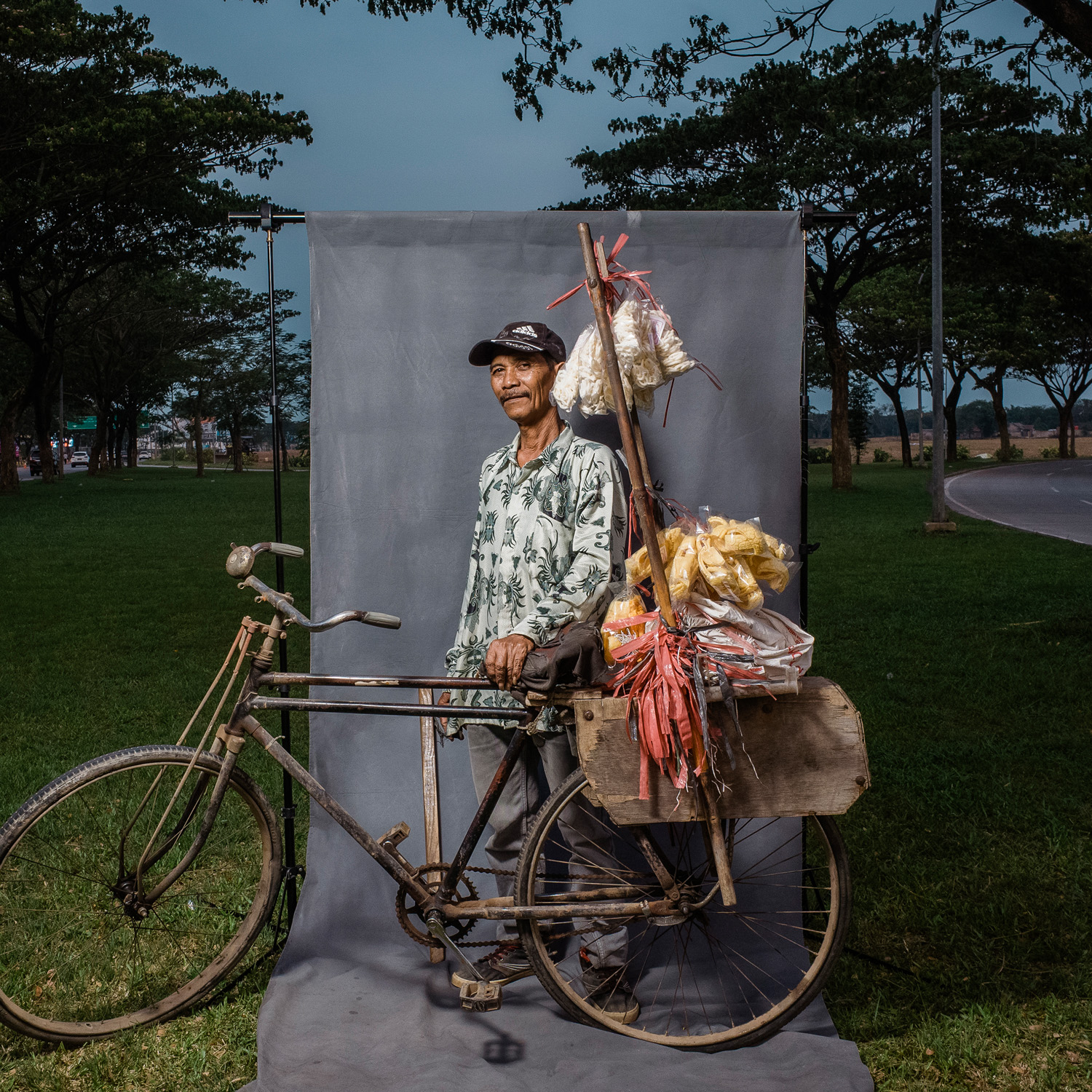 A man wearing a patterned shirt and a cap stands beside his bicycle loaded with various goods. He is posed in front of a grey backdrop in a grassy outdoor area with trees.