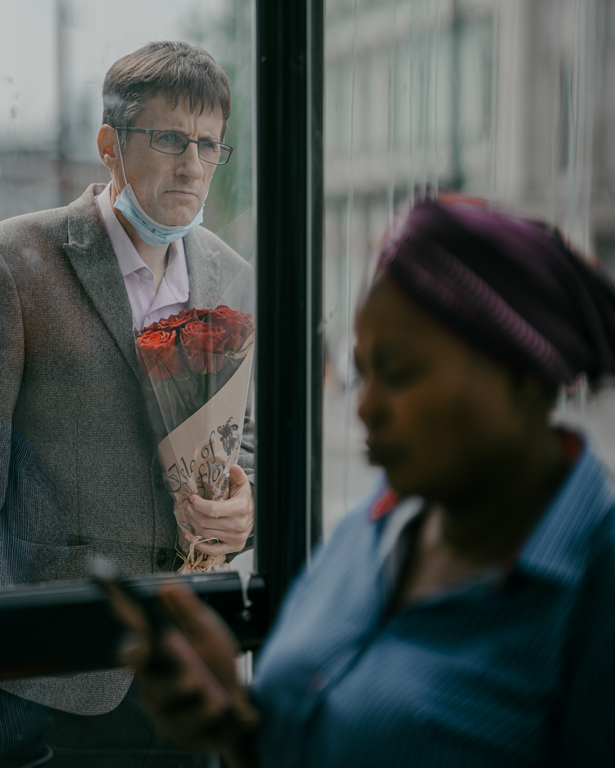 A man in a grey suit and glasses holds a bouquet of red roses and stands outside a window looking in, while a woman inside is focused on her phone.