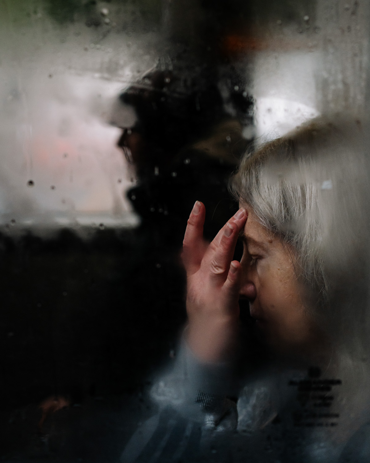 A blurred, close-up view through a foggy window showing a woman with grey hair touching her forehead, with another person in the background.