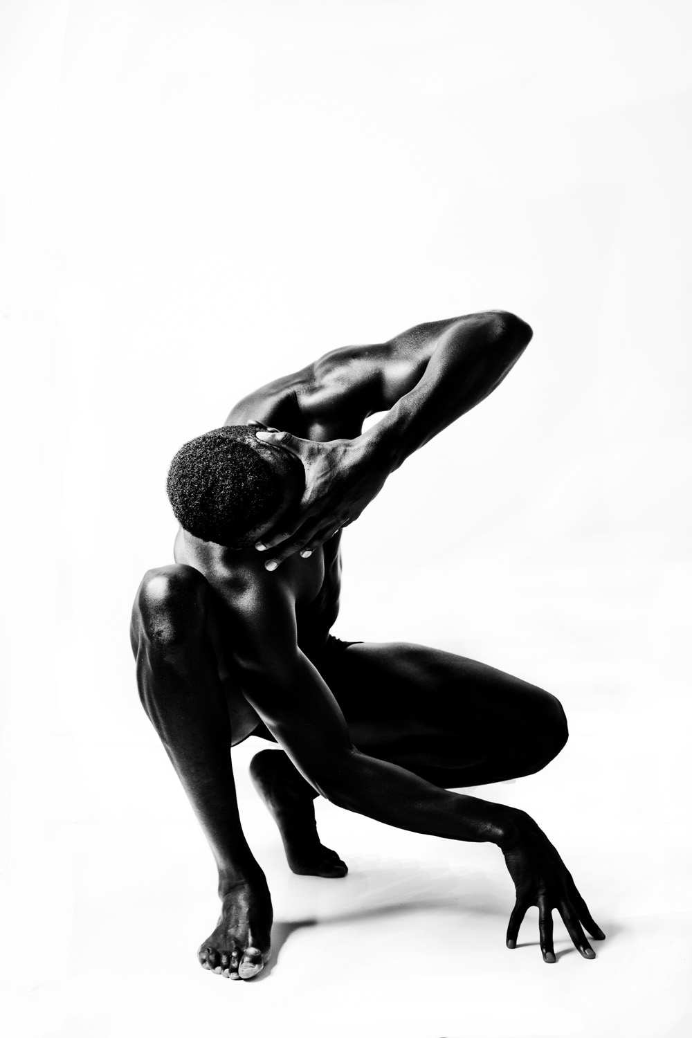 A monochrome image of a fit man in a crouched pose, one hand supporting his head and the other touching the ground, highlighting his toned physique against a stark white backdrop.