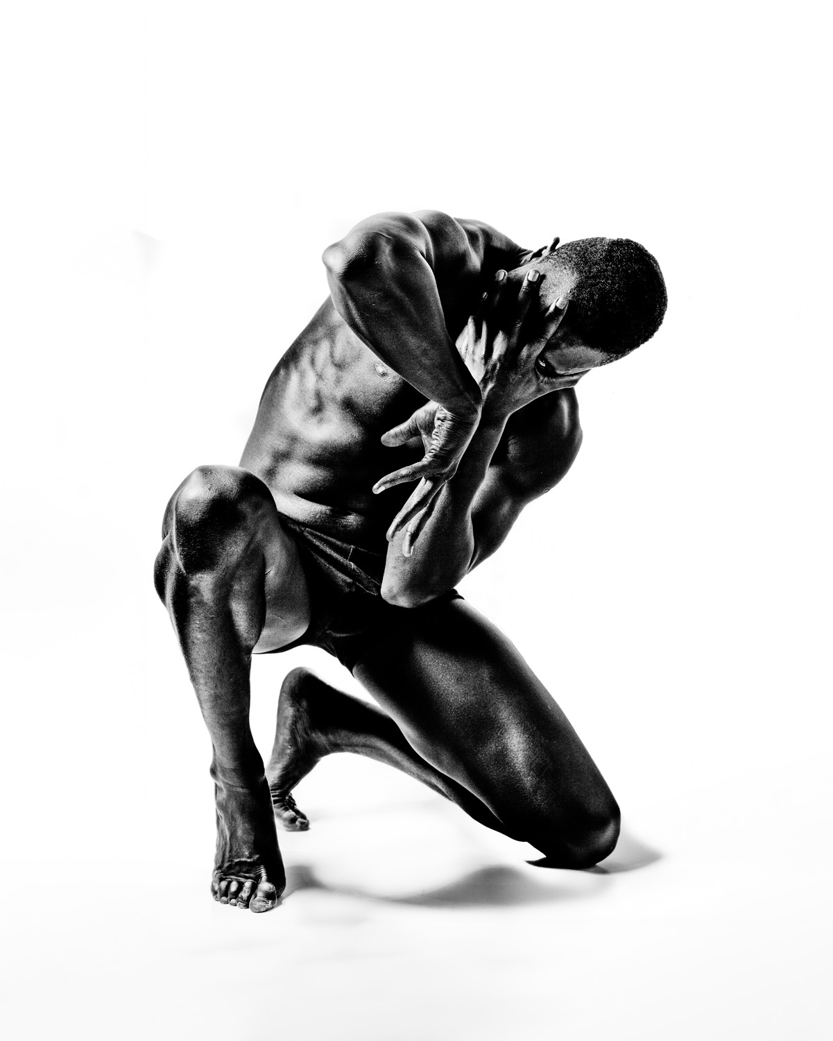 A black and white photo featuring a strong man in shorts, kneeling with one knee on the ground and hands covering his face, emphasising his muscular form and vulnerability against a white background.