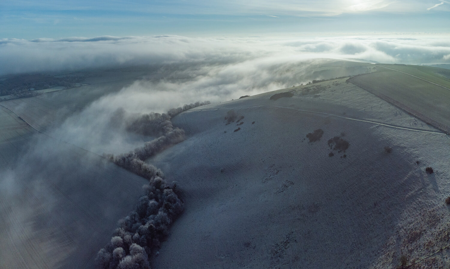 An aerial photograph capturing a rolling landscape blanketed in mist. The hills and fields are lightly dusted with frost, and a line of trees divides the scene, creating a serene and ethereal atmosphere under a clear blue sky.