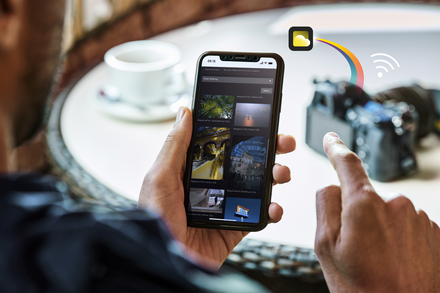 A person is using a smartphone to view and select photos. The phone screen displays various photo thumbnails of different images. In the background, a camera is placed on a table next to a cup of coffee. An icon showing a cloud with a Wi-Fi signal indicates the transfer of data from the phone to the camera.