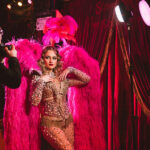 A photographer is capturing images of a model dressed in a vibrant, sparkling costume adorned with large pink feathers. An assistant is adjusting lighting equipment on the right side of the frame.