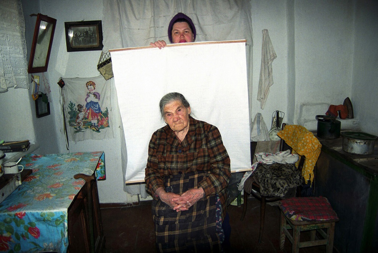 An elderly woman in a plaid dress sits on a chair with her hands clasped, looking solemn. Behind her, a person holds a white cloth as a backdrop. The setting is a modest room with various household items, including a table with a floral tablecloth and a small stove.