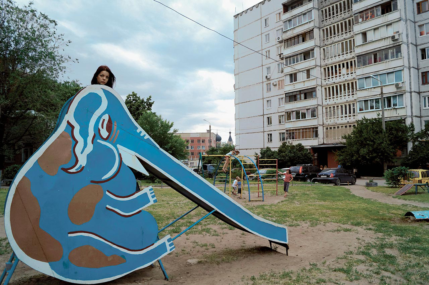 A young woman peeks over the top of a large, blue, elephant-shaped slide in a playground. The background shows an urban residential area with high-rise buildings and other playground equipment, with children playing nearby.