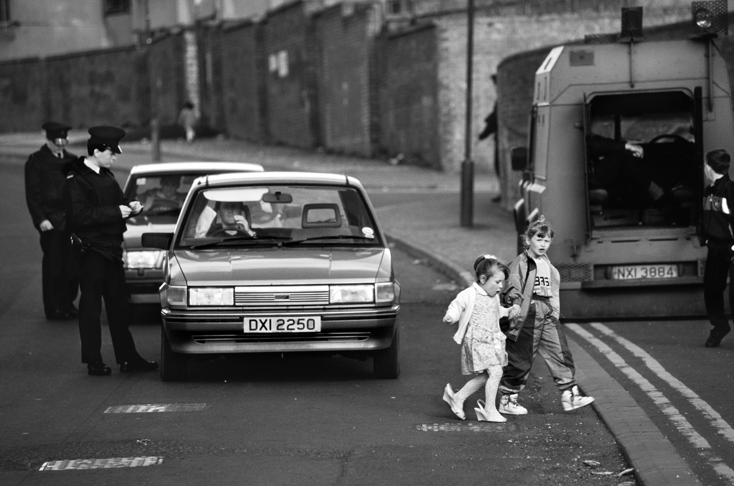 Black and white photograph of two young girls crossing a street in Northern Ireland, with police officers and armoured vehicles in the background, reflecting the everyday impact of conflict on children.