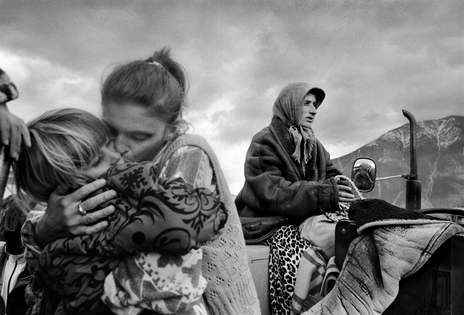 Black and white photo of a woman on a tractor holding her child close, another woman sits nearby looking into the distance, capturing the emotional toll on refugees from Kosovo.