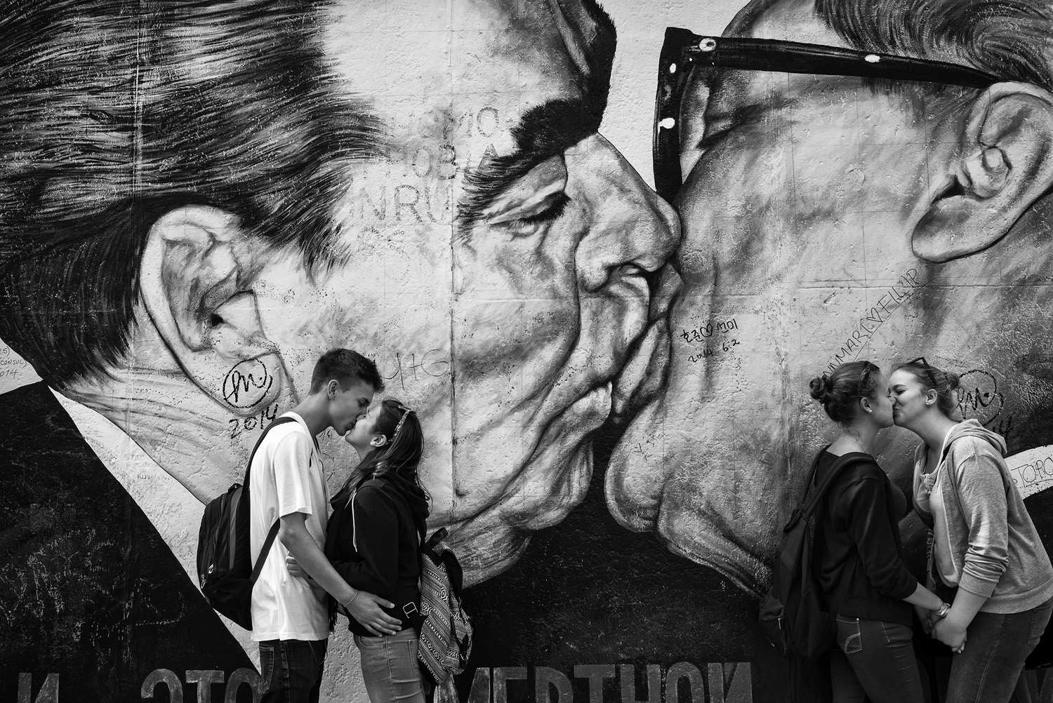 Black and white photo of couples kissing in front of a mural on the Berlin Wall depicting a famous embrace between Soviet and East German leaders, a juxtaposition of past political tension and modern love.