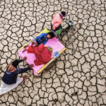 An overhead view of a man pushing a rickshaw carrying a family across a vast, cracked, drought-affected landscape, symbolising the impact of climate change on communities reliant on the land.